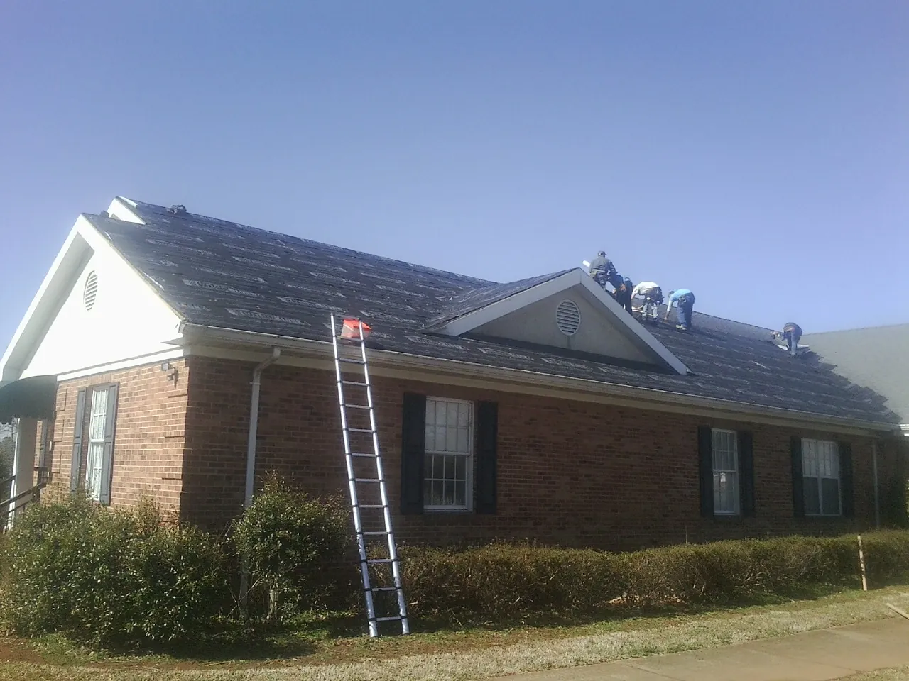 Crew tearing shingles off a brick ranch roof with a ladder set — Emergency Roof Repair work by our crew in Coinjock