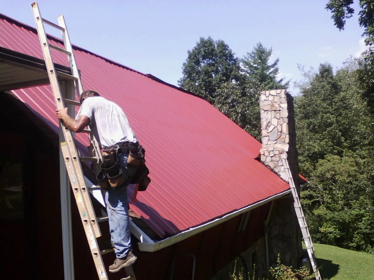 Roofer on a ladder beside a red metal roof and stone chimney — Soffit & Fascia Repair by Roofing Brothers in Coinjock